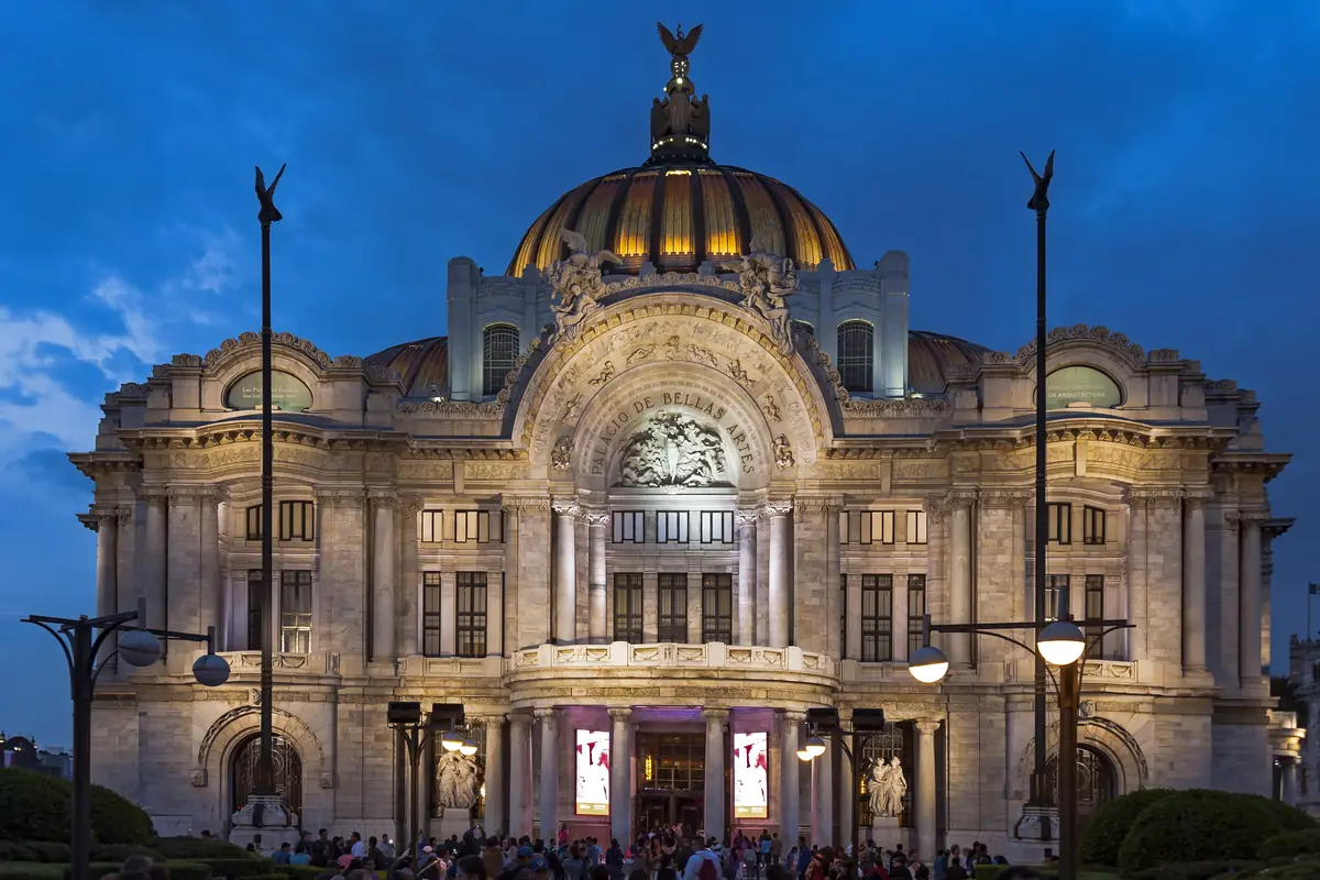 The white marble front of the Palacio de Bellas Artes lit up at dusk under a deep blue sky, with its domes, a bronze eagle on top and people gathered at the entrance