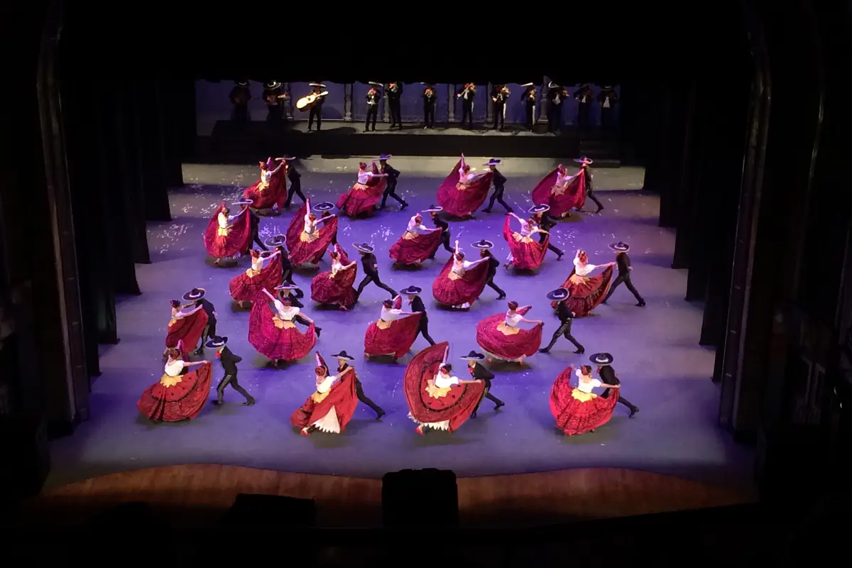 The full stage of the Palacio de Bellas Artes seen from above, with couples in red skirts and charro suits dancing and a mariachi band playing on a raised platform behind them